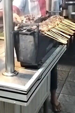 A man in a red and grey shirt is grilling satay skewers on a charcoal grill. He is standing behind a white food cart, wearing flip-flops. The skewers are arranged on top of the grill, and there is smoke rising from the charcoal. The man is looking at the food as he grills it. The surroundings are blurred, but appear to be an outdoor food stall.