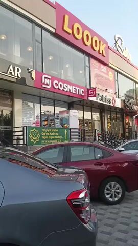 This media file captures a street view in Yangiiul, Uzbekistan, during daylight hours, possibly late afternoon given the warm lighting. The scene is dominated by the facade of a multi-tenant building. A prominent sign with large, yellow, three-dimensional letters spells out "LOOOK". Below this, a horizontal pink banner reads "M COSMETIC" with the "M" logo. To the right of the cosmetic store, another business displays "Patriss" with a similar "M" logo and a smaller sign advertising "LOOOK SALAD LONGER." Further to the right, parts of other businesses are visible, including one with "Bellissimo Pizza" and a red umbrella.

In the foreground, several cars are parked or driving by. The closest car is a dark grey sedan, partially obscuring a red sedan. Further back, a white sedan is visible.

On the sidewalk in front of the businesses, a man in a white shirt is standing, seemingly interacting with someone or observing the surroundings. A green and white sign with Uzbek text is also prominently displayed, featuring a stylized tree symbol and contact information. The overall impression is of a bustling commercial area with various retail establishments. The sky appears clear, suggesting fair weather.