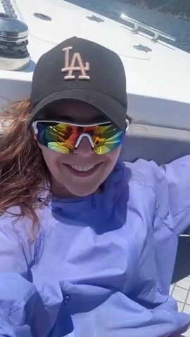 A woman with long brown hair wears a black baseball cap with a pink LA logo and mirrored wrap-around sunglasses. She is smiling and wearing a lavender hooded jacket. She is on a boat with a white deck and railing. In the background, there are blue water, distant green hills, and a blue sky with white clouds. The video is a selfie.