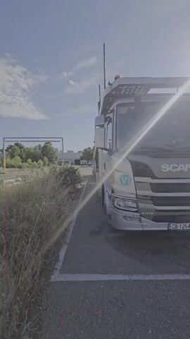 The video opens with a close-up of the front of a silver Scania truck. The camera then pans slowly to the right, revealing other trucks parked in a lot. The trucks appear to be car carriers. The environment is an outdoor parking lot under a bright, sunny sky with scattered clouds. The sun is high and bright, creating lens flare. In the background, rows of cars are visible, suggesting a car storage or distribution facility. The overall scene is static, with no visible people or actions occurring.