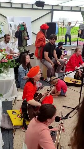 The video shows an outdoor gathering under a white canopy. In the foreground, several people are seated and playing musical instruments including guitars and drums. A woman in a red outfit is singing into a microphone. In the background, a large group of people are standing and raising their hands in unison, appearing to be participating in a group activity or celebration. The overall atmosphere seems festive and communal.