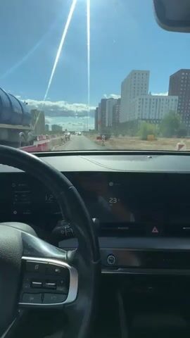The video shows the interior of a car, looking forward through the windshield. The driver's side steering wheel is visible on the left, with controls on it. The car's dashboard display shows a speed of 12.2, and the temperature outside is 23 degrees Celsius. The car is on a road under a bright, sunny sky with contrails visible. On the right side of the road, there are several tall, modern buildings. On the left side of the road, there are construction barriers and some construction equipment, including a large orange dumpster-like object. Other cars are visible on the road ahead. The overall impression is of driving through a developing urban area on a clear day.