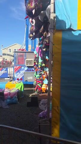 The video captures a busy outdoor carnival scene. The first frame shows a large carnival ride with blue and red structures, and a pile of large plush toys in plastic bags in the foreground. Several people are visible, including a woman holding a child. The second frame shows a man in sunglasses and a patterned shirt standing in front of a colorful carnival attraction labeled "MAZE." Other people are walking around in the background. The third frame shows a group of people near a blue spiral slide, with a woman holding a child. The fourth frame shows a man in a blue shirt and sunglasses leaning on a railing, with a carnival game booth and people in the background. The final frame shows a crowd of people in line at what appears to be a ticket booth or game area, with bright yellow signage. The overall environment is sunny with a clear blue sky, indicating fair weather.