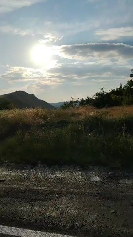 This is a short video clip filmed from a moving vehicle, likely a car, traveling along a paved road. The sun is bright and positioned high in the sky, casting a warm glow and creating lens flares. The sky is partly cloudy, with scattered white clouds against a bright blue background. The sun's rays are intense, suggesting it is midday or late afternoon.

The scene depicts a rural or semi-rural landscape. To the left, rolling hills are visible, covered in dry, golden-brown vegetation, possibly grass or brush. In the foreground, the roadside is rough and gravelly, with some scattered debris. The vehicle is moving at a moderate pace, indicated by the motion blur of the roadside and the way the landscape shifts.

To the right of the road, dense, green foliage and taller grasses are prominent. The vegetation appears somewhat wild and overgrown. In the distance, beyond the immediate roadside, more hills and possibly trees can be seen, stretching towards the horizon. The overall impression is of a journey through a sun-drenched, somewhat arid landscape. There are no people or specific objects clearly visible besides the natural elements and the road itself.

The time of day is clearly indicated by the strong sunlight and the shadows. The weather appears clear and sunny, with some clouds. The location context points to Surakhany, Azerbaijan, which is known for its oil fields and industrial heritage, but this scene appears to be a more natural, undeveloped area on the outskirts. The mood conveyed is one of movement and observation, with the bright sun and open landscape suggesting a sense of warmth and freedom. There is no visible text in the video.