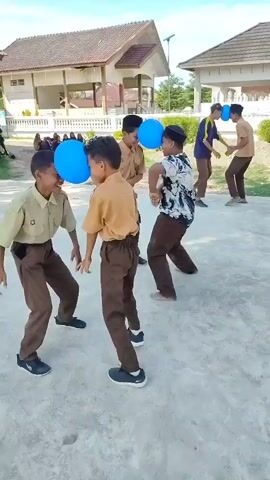 The video shows several young males, appearing to be students in uniform, participating in a game outdoors. They are holding blue balloons between their foreheads and are attempting to move forward without dropping the balloons. Some are paired up, while others are further back in the background. They are on a paved surface, with buildings and trees visible in the background. The sky is clear and blue. One person in the background appears to be filming or observing the game. The students are dressed in khaki shirts and dark brown pants. The general atmosphere appears to be lighthearted and playful.