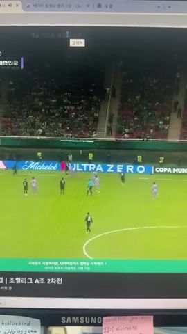 The video shows a soccer game in progress. There are several players on the field, dressed in dark and light uniforms. The field is green grass. Spectators are visible in the stands, wearing green and other colors. There are advertisements on a banner around the field. The video also shows a computer screen, with a portion of the game displayed. Text in Korean is visible on the screen.