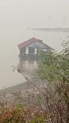 A small, unfinished wooden structure with a red roof is floating on a body of water. The structure appears to be a house or a small building, with exposed wooden beams and walls. The water level is high, with the structure partially submerged. The surrounding environment is hazy and overcast, with a shoreline visible in the foreground. The water appears muddy or murky.