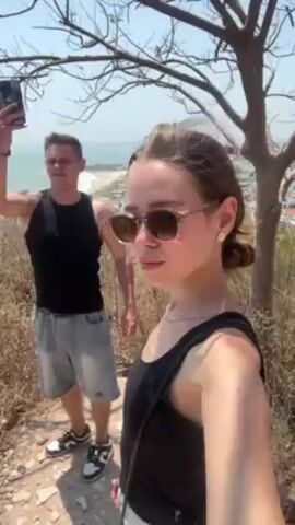 A woman in a black tank top and sunglasses is standing in the foreground on a hill.  Behind her and to the left, a man in a black tank top and jean shorts is taking a photo with his phone.  The area around them is dry, rocky, and covered in dead grass with a few bare trees nearby.  Beyond the hill, there is a beach, the ocean, and some buildings in the distance, all visible under a clear sky.