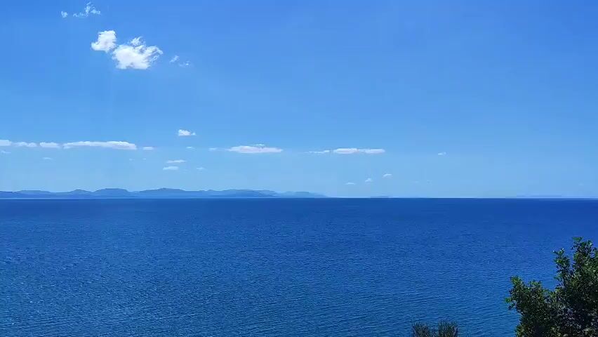 The video is a drone shot of a coastline with a shipwreck. The water is a deep blue and calm, with small waves lapping at the shore. The sky is a clear blue with a few scattered white clouds. The beach is sandy and appears to be mostly empty, with a few people visible in the distance. The shipwreck is a large, rusty vessel that appears to be partially submerged in the water. The coastline is lined with green vegetation, and in the distance, there are mountains. The drone shot pans across the scene, offering different perspectives of the shipwreck and the surrounding landscape.
