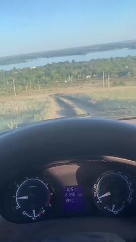 The video shows the view from inside a vehicle, looking out through the windshield. The vehicle is on a dirt road that winds through a grassy field. In the distance, there is a body of water, likely a river or lake, with a bridge crossing it. The sky is blue with some clouds. The dashboard of the vehicle is visible in the foreground, showing a speedometer, tachometer, and a digital display with the time and temperature. The overall impression is of a scenic drive through a rural landscape.
