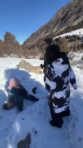 A sunny outdoor scene shows a family of three - two adults and a child - in a snowy mountainous area. The child, wearing a pink hat and dark blue snowsuit, is playing on the ground in the snow. A man in sunglasses and a green jacket points off to the side. A second woman wearing a black and white coat stands nearby. In the background, bare trees and rocky mountains are visible under a clear blue sky.