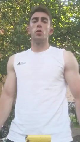 A man in a white sleeveless shirt and black shorts stands outdoors with trees and a building in the background. He pulls up his shirt to reveal his abdominal muscles.
