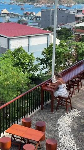 The video opens with a high-angle shot of a woman sitting at a table on a balcony overlooking a harbor. She is wearing a white dress and appears to be looking at her phone. The balcony has wooden tables and stools, and a railing. In the background, there are boats docked in the water, buildings, and lush green trees. The camera then pans across the harbor, revealing more boats and the coastline. The sky is clear and blue, and the sun is shining. The video continues to pan across the harbor, showing the bustling activity on the water and the vibrant colors of the buildings.