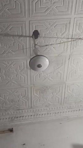 The video shows a white ceiling fan hanging from an ornate, textured, white ceiling. The fan is not spinning and appears to be partially disassembled, with the black motor housing visible at the top of the fixture. Wires hang down from the ceiling near the fan. The ceiling is made up of repeating square patterns. The bottom of the wall is visible with a decorative molding. The overall impression is one of a room in need of repair.