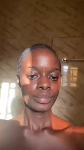 A close-up shot depicts a young woman with dark skin standing in a bathroom. She faces the camera and poses with her hand near her neck. Her hair is pulled back. Sunlight illuminates her face. The bathroom features marble-like tiled walls. A towel rack and a partially visible window are in the background.