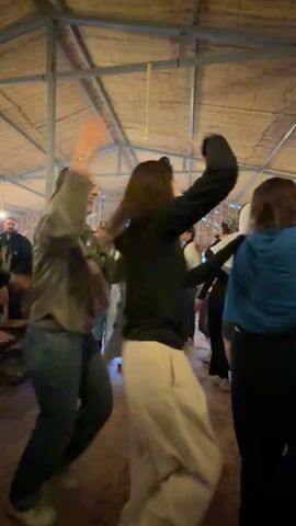 The video shows a group of people dancing together in what appears to be a large tent or open-air structure. The space has a straw or woven ceiling and a simple floor.  Several people are clapping while others dance in a circle, holding each other's hands or shoulders. The group is diverse in terms of age and attire. One person is wearing traditional Middle Eastern clothing. There is a fire pit visible in the background, and other people are seen watching and recording. The atmosphere seems lively and festive.
