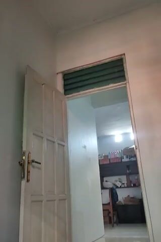 Here is a detailed description of the video:

**Scene:** The video presents an interior shot of a room, focusing on a doorway or opening that leads to another room.

**Environment:** The walls are painted white. The lighting appears to be bright, possibly from an overhead light source in the adjacent room.

**Objects:**
*   **Door:** A white, paneled door hangs open. It's attached to the top frame and swings downwards into the room where the video is being shot. The door has a simple locking mechanism in brass or gold color.
*   **Window:** To the side of the door opening, there's a window with green blinds.
*   **Adjacent Room:** Through the doorway, the adjacent room appears brighter. There's a bookshelf filled with items, some boxes, and what looks like a television.

**Camera Work:** The camera is held relatively still, but there is some slight shakiness. It's pointed upwards, capturing the doorway and the ceiling. The camera angle suggests that the video was taken from a relatively low perspective.

**Overall Impression:** The video gives a glimpse into a simple, functional space with slightly disheveled or unfinished aspects.