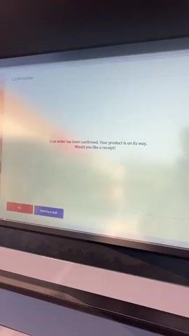 The video shows a close-up of a touchscreen display. The screen displays a confirmation message stating "Your order has been confirmed. Your product is on its way. Would you like a receipt?". Below this, there are two buttons: a red button labeled "No" and a purple button labeled "Send by e-mail". Further down the screen, in French, it says "Ton avis est précieux pour la famiglia" which translates to "Your opinion is precious to the family". There are two green buttons below this French text, one labeled "Je réponds !" (I answer!) and the other "Je passe mon tour." (I pass my turn.). Below that, it states "Product(s) remaining to be delivered: 1/1" and then in red text, "Your Margarita is on its way...". The screen has a soft, blurry reflection of a person and the surrounding environment, suggesting it's a public terminal or kiosk. The lighting is somewhat dim, and the focus is primarily on the screen content.