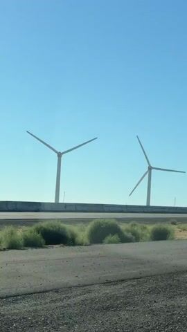 This video shows a view of wind turbines in a desert landscape. The turbines are tall, white structures with three blades each. They are positioned along a highway overpass, with sparse vegetation and dry ground visible below. The sky is a clear, bright blue, suggesting a sunny day with good visibility. The camera appears to be mounted in a moving vehicle, as evidenced by the slight movement of the scene and the blurry foreground. There are no people visible in the video. The overall impression is one of open space and industrial energy generation in a somewhat arid environment. The time of day appears to be daytime, likely mid-morning or afternoon, given the brightness of the sky and the shadows cast. The location context of Tracy, United States, is consistent with a desert or semi-arid landscape with wind farms.