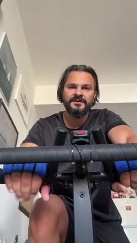 A man with dark hair and a beard, wearing a black t-shirt and black shorts, is riding a stationary bicycle indoors. He has blue fingerless gloves on and is holding the handlebars. The bicycle has a black frame and handlebars wrapped in black tape with blue grips. There is a red indicator light on the handlebars. The background shows a wall with several framed pictures and a white ceiling. The lighting appears to be natural daylight. The man is looking directly at the camera and appears to be speaking.