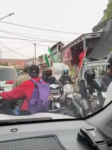 The video shows a street scene, likely in Southeast Asia, with a traffic jam of motorcycles and a car. Several people are visible on motorcycles, some wearing helmets and carrying backpacks. Green and red flags are displayed on some of the motorcycles and in the background. The environment appears to be a residential or commercial area with buildings lining the street and overhead power lines. The perspective is from inside a vehicle, looking out at the traffic. The overall atmosphere seems to be one of congestion and possibly a procession or gathering.