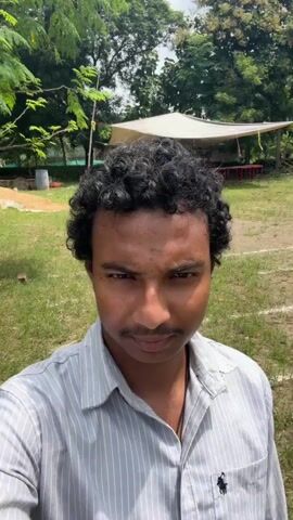 A person with dark curly hair and a mustache is taking a selfie outdoors. They are wearing a light grey striped button-down shirt. In the background, there are trees, grass, and what appears to be a field with white lines marking a track or boundary. There are also some structures and other people visible in the distance. The person smiles at the camera and holds up two fingers in a peace sign.