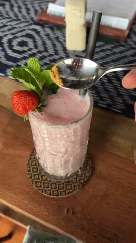 The video shows a pink strawberry smoothie in a textured glass on a woven coaster, sitting on a wooden table with a blue patterned tablecloth in the background. A metal straw is in the smoothie, and it's garnished with a fresh strawberry and a sprig of mint. Initially, a hand sprinkles some golden crumbs onto the top of the smoothie from a spoon. Then, another person pours a stream of the pink smoothie into the glass, presumably from a blender or another container. The overall scene suggests a close-up shot of a refreshing beverage being prepared or served in a cafe or restaurant setting.
