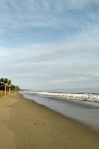 The video shows a long, sandy beach with gentle waves rolling onto the shore. The sky is a mix of blue and white clouds, giving a bright but slightly overcast feel to the scene. On the left side of the frame, there is some greenery with what appears to be a small structure partially visible. The beach is mostly empty, with only a few scattered objects visible on the sand. The camera seems to be stationary, providing a wide view of the beach and the sea.