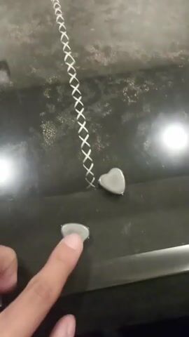 A person's left hand is visible in the lower portion of the frame, with the index finger touching a small, heart-shaped object. To the right of this object is another identical heart-shaped object. Behind these objects is a dark surface with a white criss-cross pattern. The surface appears to be textured and possibly metallic or stone. A bright light source is reflected in the upper right quadrant of the frame. The lighting is dim overall, with the primary light source coming from the right.