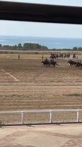 The video shows a group of jockeys racing horses across a dirt track, with a body of water visible in the background. The view is from a seated position looking through a barrier.