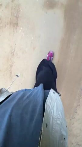 The video begins with a close-up view of a person's legs and feet walking on a dirt path. The person is wearing dark pants and pink and black athletic shoes. The camera then pans up and moves forward, revealing a wide dirt road flanked by dense green trees. In the distance, a dog, appearing to be a husky or malamute with black and white fur, is running towards the camera on the road. The dog is moving quickly and its tail is held high. The scene appears to be outdoors in a natural, possibly wooded, environment. The lighting suggests it is daytime.