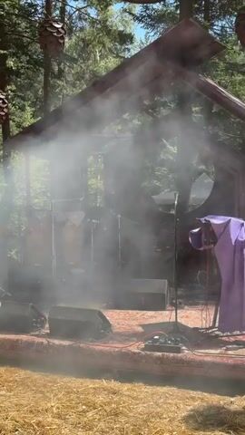 The video shows an outdoor stage set up in a wooded area. There is a structure resembling a small house with a roof, and trees are visible in the background. Smoke or fog is present on the stage, obscuring some of the equipment. On the stage, there are musical instruments, including drums and possibly a keyboard or DJ setup. A microphone stand is also visible. A purple cloth is draped over an object on the right side of the stage. In the background, a person is partially visible behind the smoke, appearing to be operating some equipment. The ground in front of the stage is covered with straw or hay. There are also some cone-shaped decorations hanging from above.