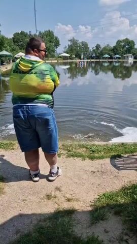 A person is fishing at the edge of a lake. The person is wearing a green and yellow hooded jacket with the text "SeaBuzz" on the back, denim shorts, and white sneakers. They are holding a fishing rod, and the line is in the water, creating ripples. There is foam on the water's surface near the person.

The scene is outdoors with trees in the background and a clear blue sky with some clouds, suggesting it is daytime and the weather is fair. Across the lake, there are several white umbrellas or canopies, possibly indicating a recreational area or a fishing spot with amenities. In the distance, there are some colorful cylindrical objects on the water. The ground in the foreground is a mix of dirt and grass. The overall impression is a sunny day at a fishing location.

There is no explicit mention of German or Bulgarian context in the visual information provided.