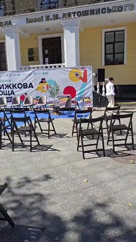 The video shows an outdoor gathering in front of a yellow building with a sign that reads "МУЗИЧНА ШКОЛА імені Б.М. ЛЯТОШИНСЬКОГО". In front of the building is a white banner with colorful graphics and text, including "КНИЖКОВА ПЛОЩА". There are several black folding chairs set up on a grey paved area, facing the banner. Some chairs are empty, while others have people sitting in them. In the foreground, three people are visible sitting in chairs, facing away from the camera. A woman in a white shirt and dark pants walks from right to left behind the banner. The lighting suggests it is daytime.