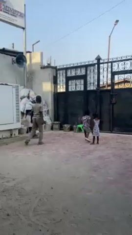 A few children and an adult are in an open, paved area near a building that has a sign, "FIRE MINISTRY SOULS CHURCH", on the left of the frame. Next to the building, there is a large white object covered with bags of material. In the center of the scene, a man in uniform passes a soccer ball to a boy. Another boy stands nearby with the ball in his hands. Two other kids stand to the right near a closed metal gate. A green chair is visible to the right. The ground has spots of red or pink color. The sky is blue.