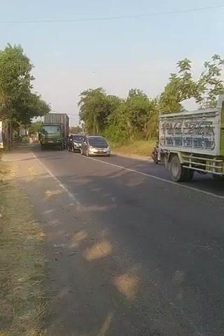 A green truck with a large shipping container is on a two-lane road, moving away from the camera. To the left of the truck, two cars are stopped, one silver and one black. To the right of the truck, a motorcycle carrying one person is moving in the same direction as the truck. Further down the road to the right, a group of motorcycles carrying multiple people are also moving in the same direction. Trees and vegetation line both sides of the road, and the sky is clear. The scene appears to be in a rural or suburban area during daylight.