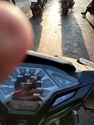The video begins with a close-up of a motorcycle's dashboard, displaying a speedometer with the needle at approximately 0 km/h and an odometer reading of 88902.9 km. The fuel gauge appears to be low. The perspective then shifts to a first-person view from the motorcycle, showing a road with several other motorcycles ahead, forming a convoy. The sun is low in the sky, casting long shadows across the asphalt. The road appears somewhat worn and uneven. The other motorcyclists are mostly wearing helmets, and some are carrying passengers. The environment suggests an urban or suburban setting with buildings and trees visible along the side of the road. The video captures the group of motorcycles traveling down the road, with the sun creating a warm, bright atmosphere.