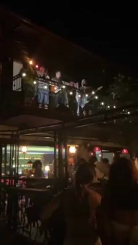 The video shows an outdoor restaurant scene at night. A band of four people is playing on a balcony or elevated platform, using a microphone and at least two guitars. The restaurant below is dimly lit with string lights and hanging lamps. People are seated at tables and standing around, mostly in the shadows, but it appears that they are watching and listening to the music. The overall atmosphere is lively and relaxed.
