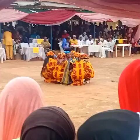 The video captures an outdoor cultural performance. In the foreground, the backs of three individuals wearing headscarves (pink, black, and red) are visible, facing away from the camera. In the midground, a group of approximately 6-8 individuals, dressed in vibrant orange, yellow, and black patterned traditional attire with matching head coverings, are performing a synchronized dance while crouched in a tight circular formation on a dirt ground. They move their upper bodies and arms subtly. In the background, numerous spectators are seated at white-clothed tables under a large canopy tent draped with red, white, and green fabric, watching the performance. Some audience members are standing.