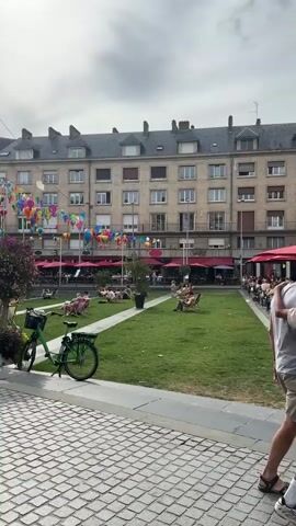 The video pans across a public square decorated with numerous colorful hot air balloon-shaped ornaments suspended from wires between tall poles. The square features a manicured lawn where several people are relaxing in lounge chairs, some appearing to be reading or resting. A green bicycle is parked on a cobblestone path in the foreground. In the background, a multi-story building with numerous windows lines the square, and outdoor seating areas with red umbrellas suggest a cafe or restaurant. A small water feature is visible on the lawn, and a person is walking near it. The sky is overcast.