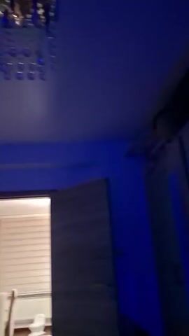 The video pans across a room with blue lighting. On the left wall is a round decorative clock. To the right of that, a doorway is visible, leading into another room with blinds on the window. At the top of the frame, a modern chandelier with clear crystal elements hangs from the ceiling.