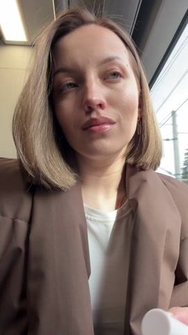 A young woman with short blonde hair and wearing a brown jacket is on a train. She unscrews a small container of lip gloss with a white cap and applicator. She applies the gloss to her bottom lip and then looks directly at the camera with a slight smile. The train window is visible in the background.