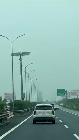 A white SUV is driving down a highway. The sky is overcast and the road is wet, suggesting it is raining or has recently rained. The highway is lined with streetlights and signs, and there are other vehicles on the road. The camera is positioned behind the SUV, capturing the view from the back seat. The video is a continuous shot with no cuts.