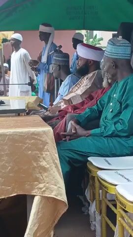 Here is a description of the video:

The video shows a group of men seated under a green tent, seemingly attending an event. They are dressed in traditional attire, including robes and hats, and are seated at a table covered with a gold cloth. Some are holding phones or have them in their laps. Chairs with "OKOBRONNU" printed on them are provided for seating. The atmosphere seems formal, suggesting a meeting or ceremony of some sort.