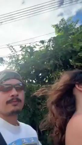 The video, shot from a moving vehicle, shows two people riding along a narrow road. The person filming is on the driver's side, wearing a camo bandanna and sunglasses. A woman with long curly brown hair is beside them. The road is paved and has a yellow line marking the center. Lush green foliage lines the road on both sides. Utility poles and wires are visible against the sky. The sky is partially cloudy with patches of blue. In the distance, another vehicle is visible on the road. The overall environment suggests a rural or suburban setting.