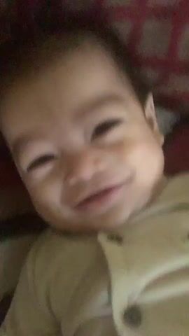 This is a close-up video of a baby with dark hair and eyes. The baby is wearing a light-colored shirt with buttons. The baby is smiling and appears to be interacting with someone off-camera. The background is blurry and appears to be a patterned surface, possibly a blanket or rug. The lighting is dim. The video is short, lasting only a few seconds, and the baby's expressions change slightly throughout.