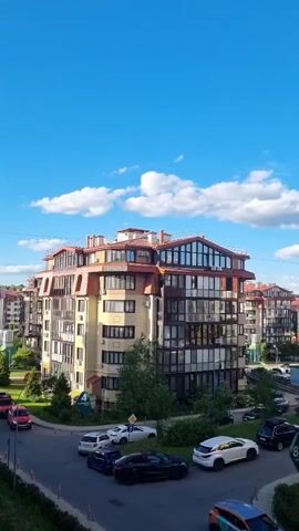 This is a vertical video capturing a view of a modern residential complex on a clear, sunny day. The buildings are multi-story with light-colored facades and distinctive terracotta-tiled, mansard-style roofs. Many have large windows and balconies, suggesting contemporary architecture. The scene is set in an urban or suburban environment with well-maintained greenery, including trees and bushes lining the streets and surrounding the buildings.

Several cars are visible parked on the street and in what appears to be a driveway or designated parking area. The cars are of various makes and models, and include a red SUV, white sedans, a dark sedan, and a dark SUV. The street itself is paved and curves through the complex.

The sky is a vibrant blue, dotted with fluffy white clouds, indicating fair weather. The sunlight casts shadows, suggesting it is daytime, likely late morning or afternoon. The overall impression is one of a peaceful and well-ordered residential area.

There are a few signs visible:
- A blue and white sign with a number '4' on a green background.
- A green sign with white text that appears to read "8к2".
- A blue directional arrow sign.

There are no people visible in the frame. The video appears to be taken from an elevated perspective, perhaps a balcony or window of another building within the complex. The setting is likely Romashkovo, Russia, as per the location context. The mood is calm and serene, reflecting the pleasant weather and quiet atmosphere.