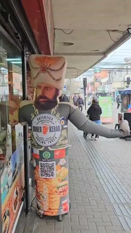 The video shows a Turkish restaurant advertisement on a sidewalk. The advertisement is an inflatable, cylindrical figure with a picture of a bearded man in a tall, ornate hat on top. The figure has arms extended outward. Printed on the body of the figure is "Autentica Doner Kebab" and "Cocina Turca" with a QR code for the menu, and images of food. The figure is positioned next to the restaurant's front window. Pedestrians walk by on the sidewalk in the background. The street includes other buildings and street furniture. The day appears overcast.