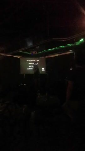 A young woman stands in a dimly lit room, possibly a bar or karaoke venue, singing into a microphone. A projector screen behind her displays song lyrics. The screen shows the text "Her boyfriend's a d**k" and "And he brings a gun to school". Green LED lights are visible above the screen. The woman is wearing a white shirt and a dark skirt with suspenders. A table with a microphone stand is in front of her. Bottles of drinks are visible on a surface in the foreground. The room is mostly dark, with the screen and lights providing the main illumination. Another person is partially visible to the right of the frame.