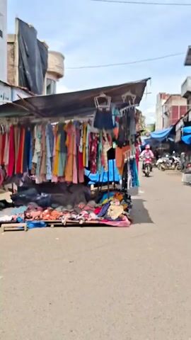 The video shows an outdoor market stall with clothing for sale. The stall has a dark tarp as a roof and is lined with racks of colorful garments, including dresses, shirts, and skirts. At the bottom of the stall, piles of clothing, including what appear to be undergarments, are laid out on a pink mat. To the right of the stall, a narrow street is visible with other stalls and buildings in the background. A person on a motorcycle, wearing a pink helmet and jacket, rides down the street. Another person, wearing a white shirt and a sarong, walks further down the street away from the camera. The weather appears to be sunny with some clouds in the sky.